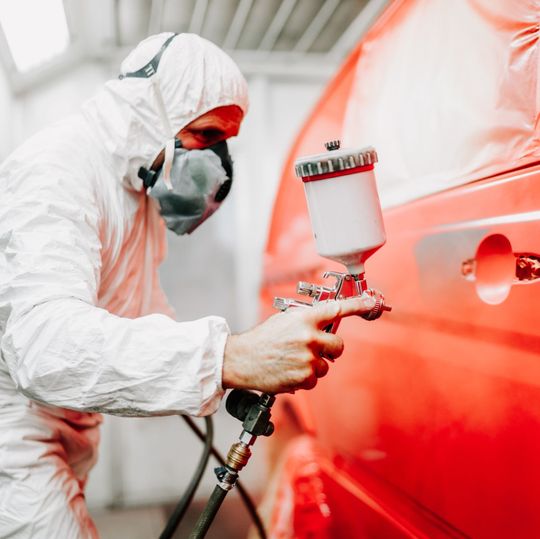 trabajador de taller con traje y mascarilla pintando de rojo una furgoneta